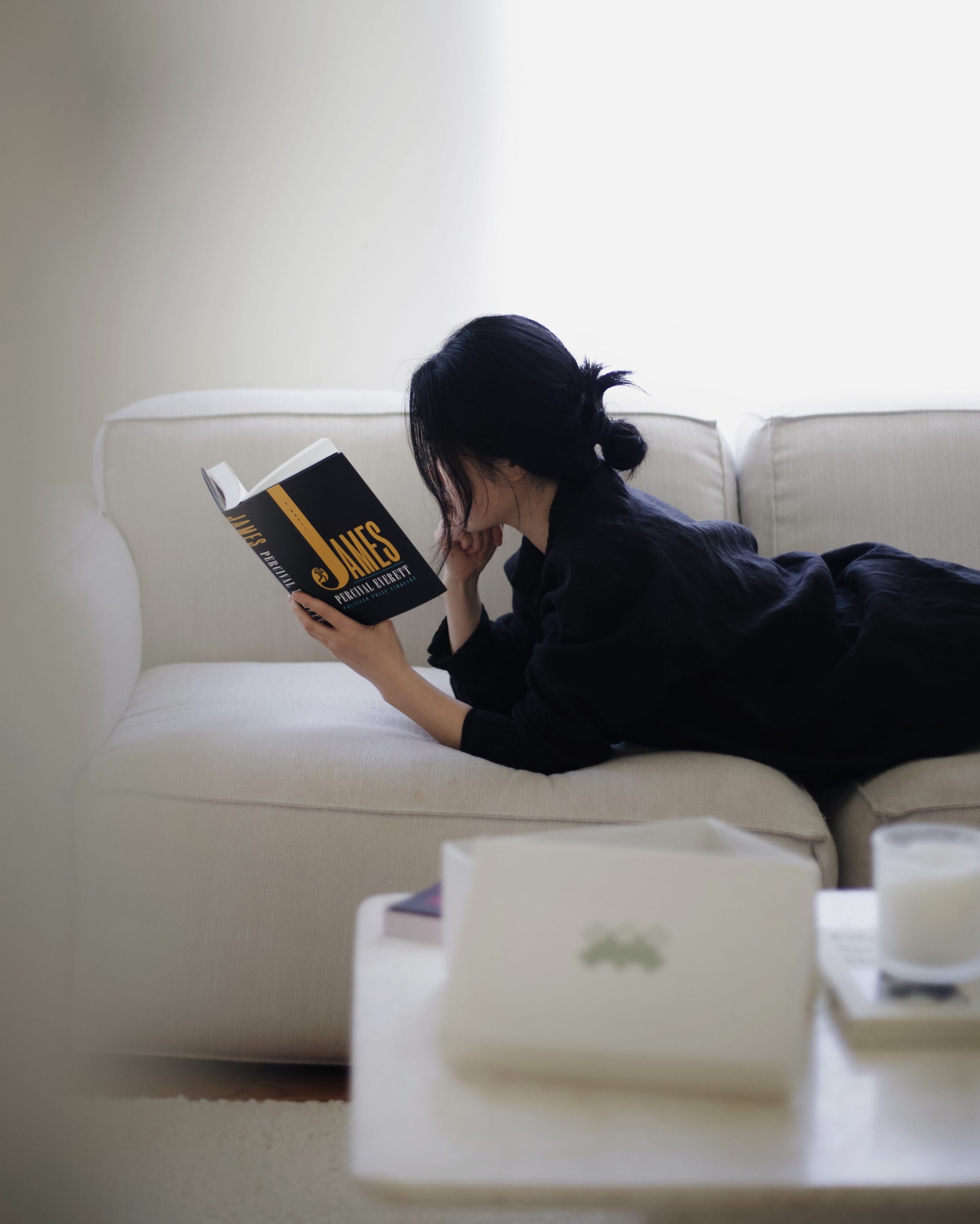 Image of woman in black clothes lying on a white couch and reading James by Percival Everett